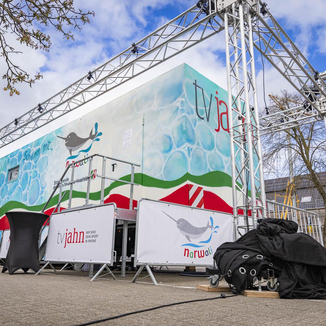 Bauger&uuml;st mit beleuchtetem Podium und Werbung an einer Wand, umgeben von Schutzh&uuml;llen auf einem Platz im Freien.