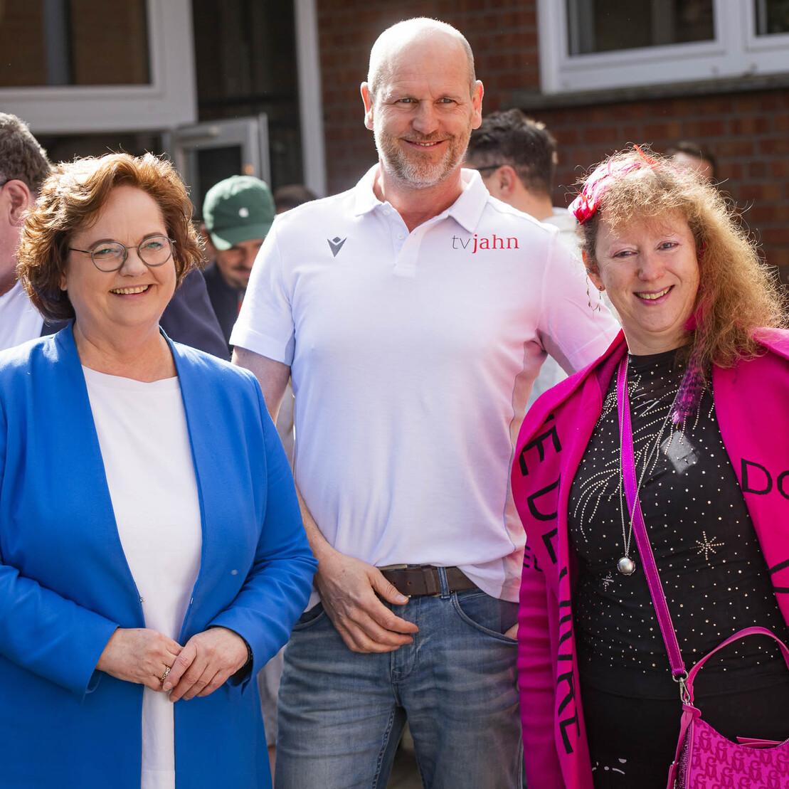 Drei Personen stehen l&auml;chelnd vor einer Gruppe von Menschen in einem Garten, umgeben von gr&uuml;nen Pflanzen und Geb&auml;uden.