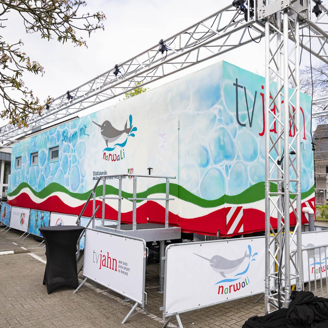 Mobiles Veranstaltungsgeb&auml;ude mit buntem Design und Absperrung auf einem Parkplatz, umgeben von B&auml;umen und Geb&auml;uden.