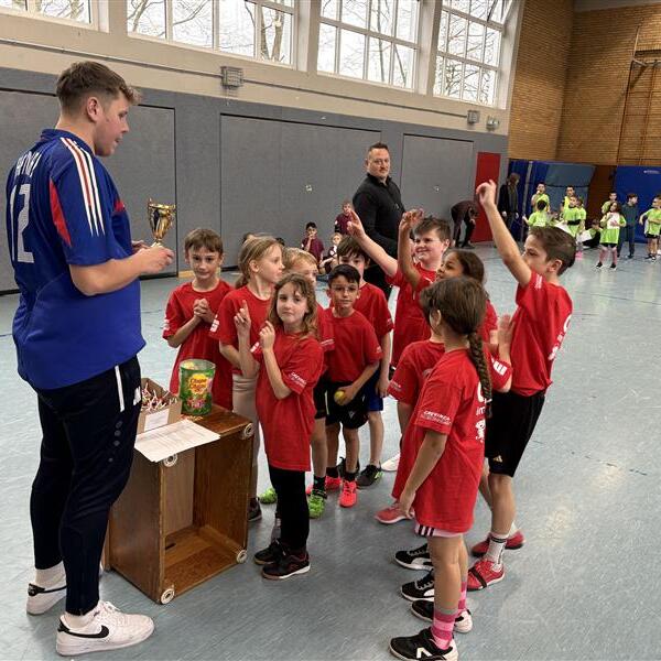 Kinder in roten T-Shirts freuen sich um einen Trainer, der einen Pokal in einer Sporthalle pr&auml;sentiert.