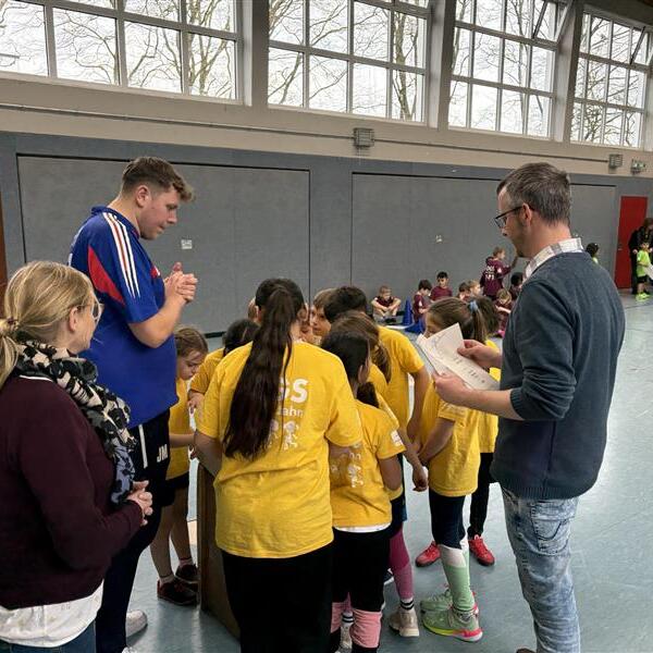 Gruppendiskussion von Kindern in gelben Trikots, geleitet von zwei Erwachsenen in einer Sporthalle.