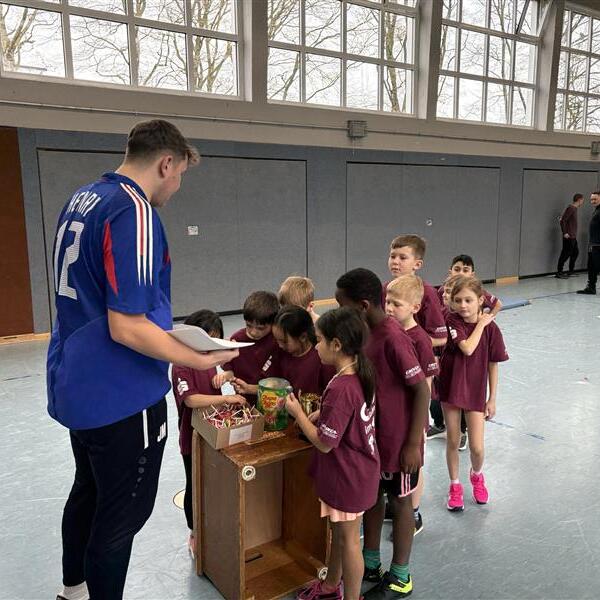 Trainer steht vor einer Gruppe von Kindern in Sportkleidung, die an einem Tisch mit Materialien stehen.