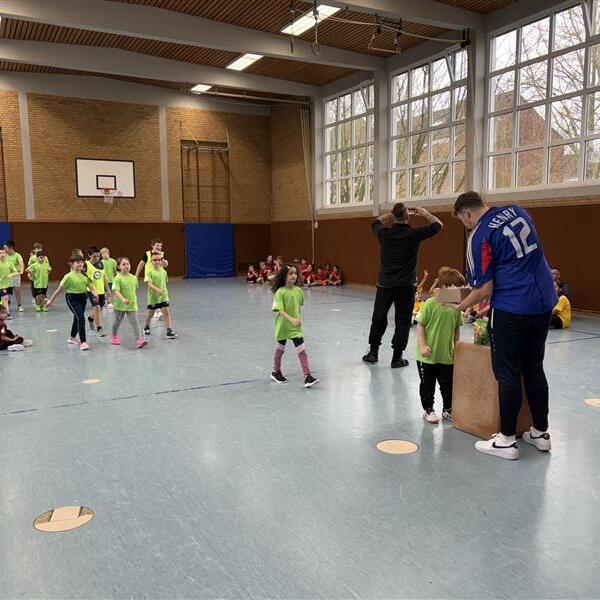 Kinder in gr&uuml;nen T-Shirts stehen in einer Sporthalle, w&auml;hrend Betreuer an einer Kiste mit Preisen arbeiten.