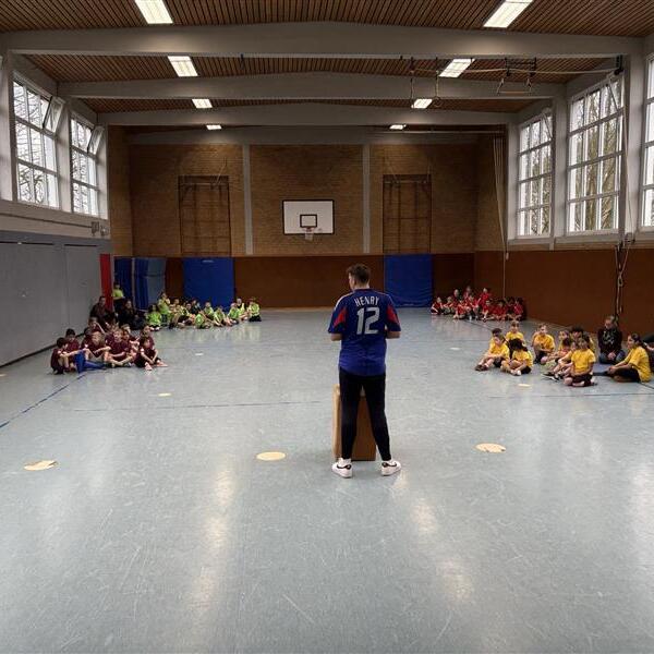 Junge im blauen Trikot spricht vor Gruppen von Kindern in bunten Sportanz&uuml;gen in einer Turnhalle.