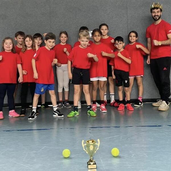 Gruppe von Kindern in roten T-Shirts, l&auml;chelnd, vor einer Wand mit einem Pokal und zwei Tennisb&auml;llen auf dem Boden.