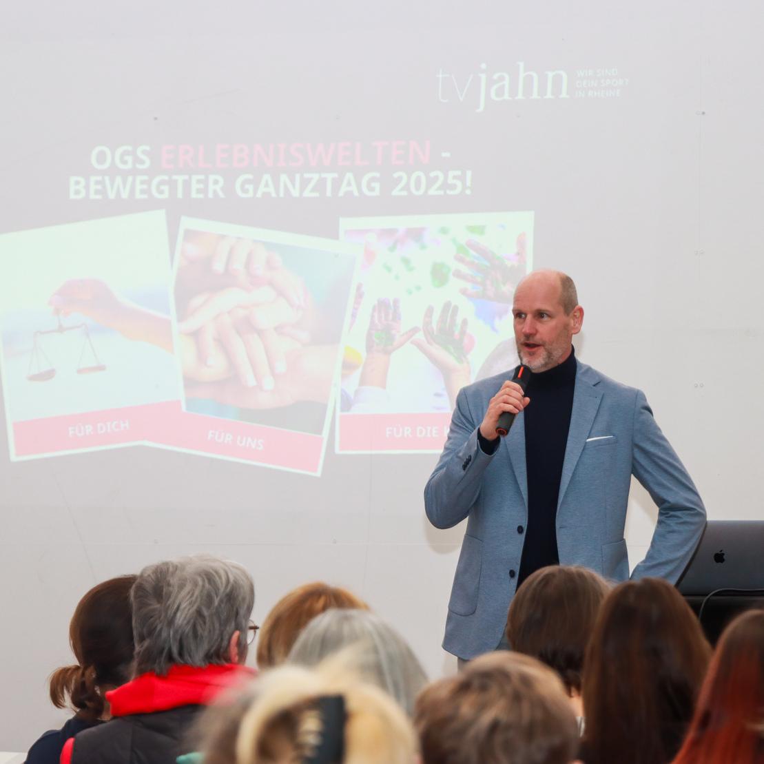 Mann spricht auf einer Veranstaltung vor Publikum, Pr&auml;sentation mit dem Titel &bdquo;OGS Erlebniswelten &ndash; Bewegter Ganztag 2025!&ldquo; im Hintergrund.