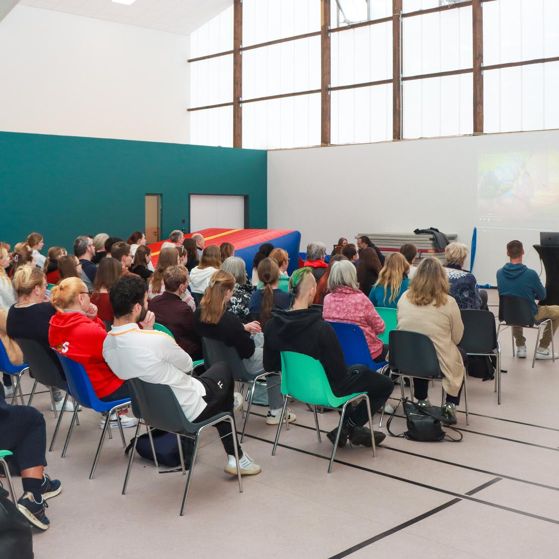 Gruppe von Menschen sitzt in einem Raum, w&auml;hrend eine Person auf einer B&uuml;hne eine Pr&auml;sentation h&auml;lt.