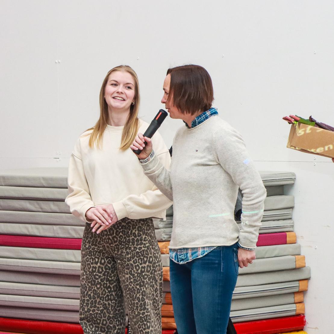 Zwei Frauen in einem Sportraum: Eine spricht mit Mikrofon, die andere l&auml;chelt. Im Hintergrund sind Turnmatten sichtbar.