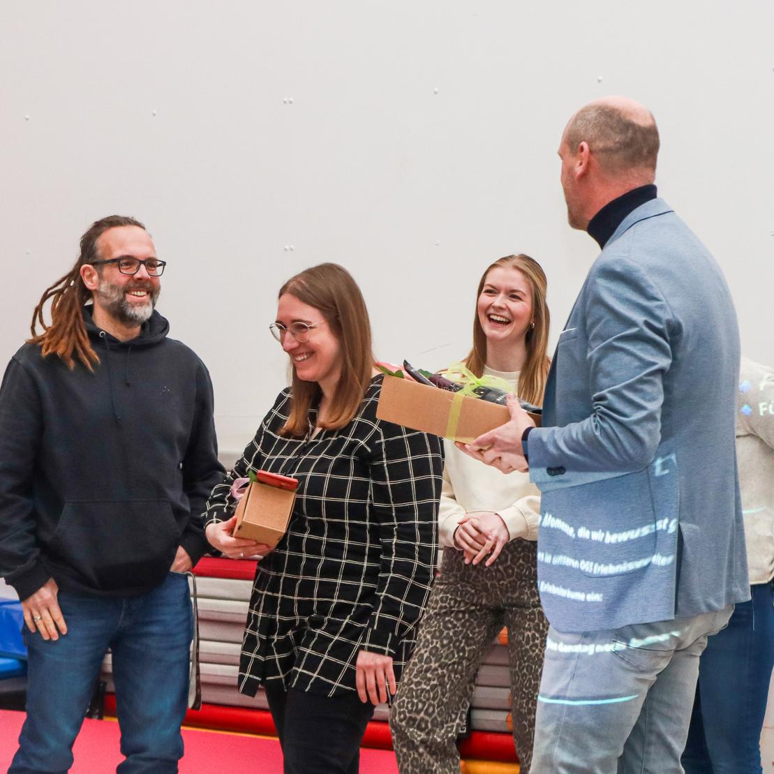 Vier Personen in sportlicher Umgebung tauschen Geschenke aus; sie lachen und freuen sich gegenseitig.