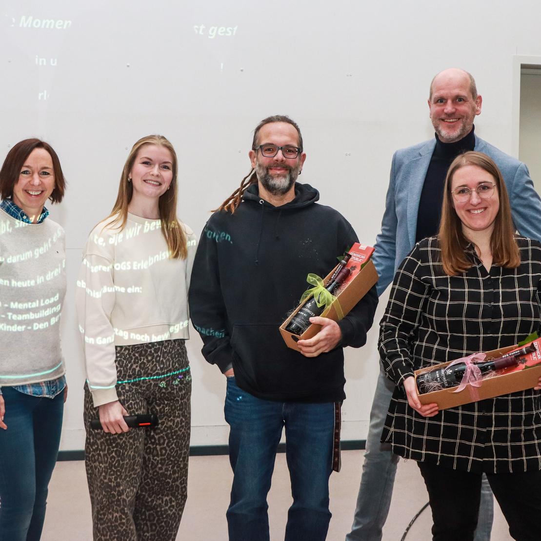 F&uuml;nf Personen stehen in einer Gruppe, halten Geschenkk&ouml;rbe und l&auml;cheln vor einem hellen Hintergrund.