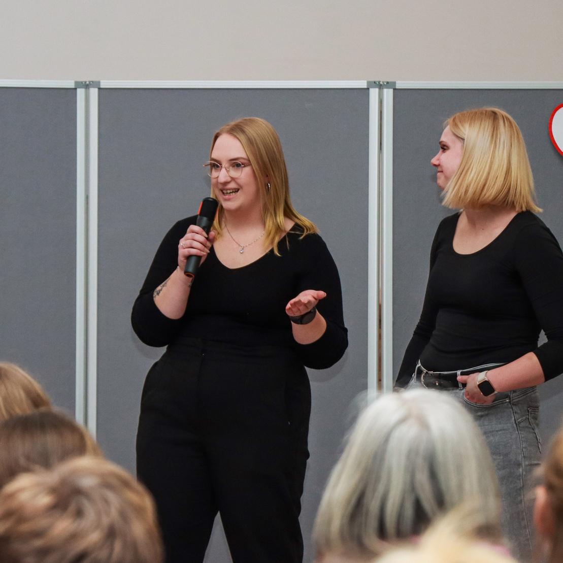 Zwei Frauen stehen bei einer Pr&auml;sentation vor einem Publikum, eine mit Mikrofon, die andere beobachtend.