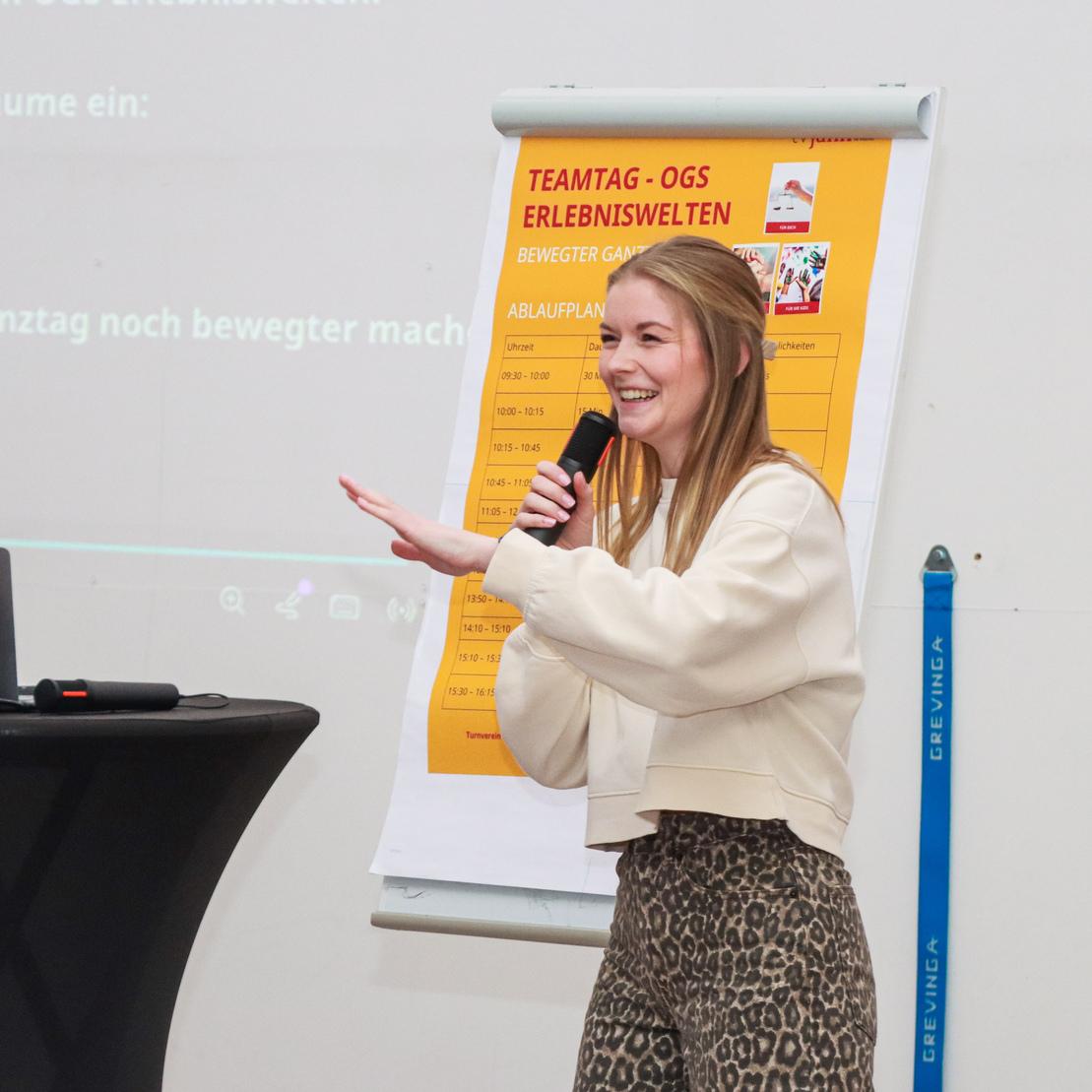 Frau spricht mit einem Mikrofon vor einem Flipchart und einem Laptop w&auml;hrend einer Veranstaltung.