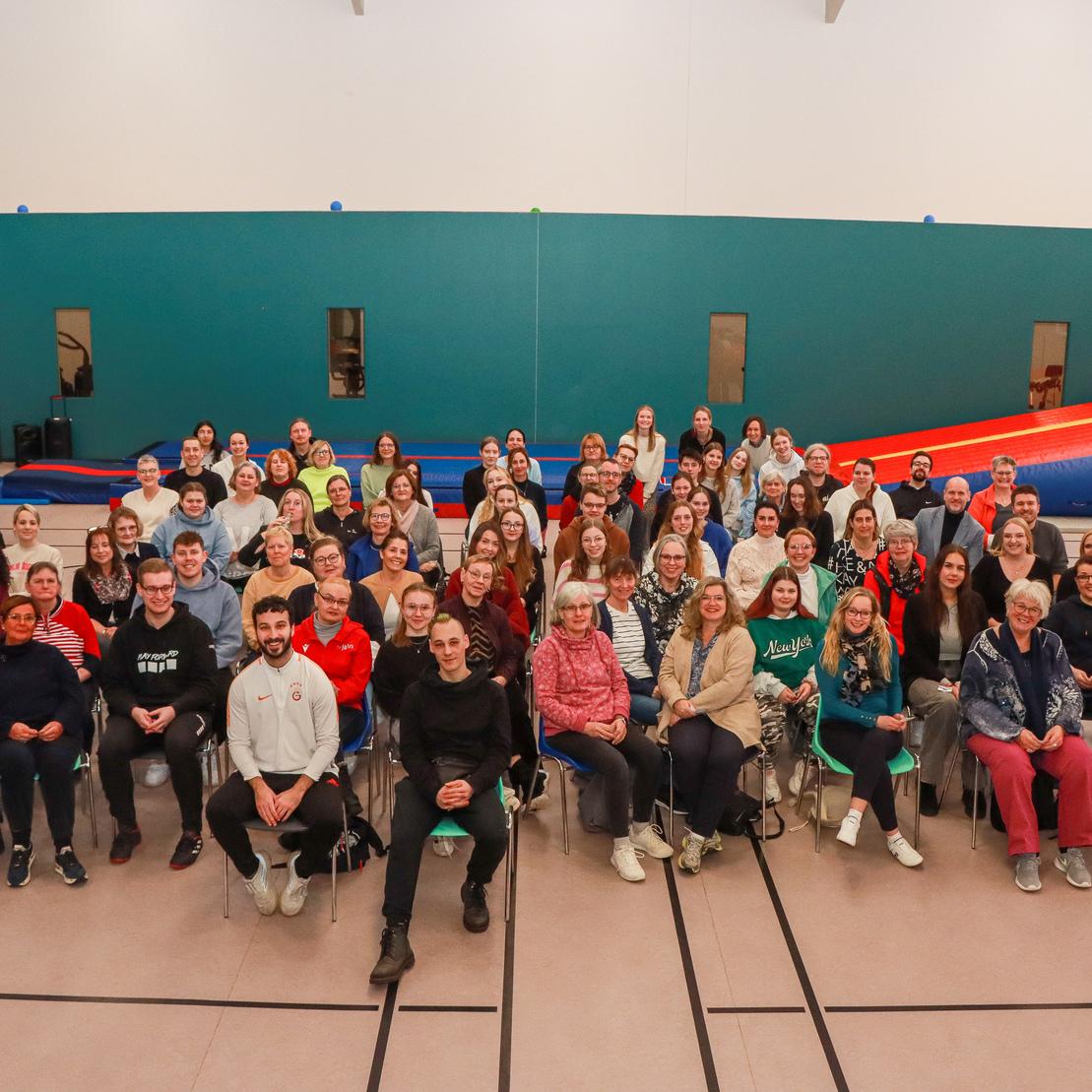 Gruppenbild von &uuml;ber 100 Personen in einem Sportraum, umgeben von Turnger&auml;ten und Matten, mit freundlichen Gesichtern.