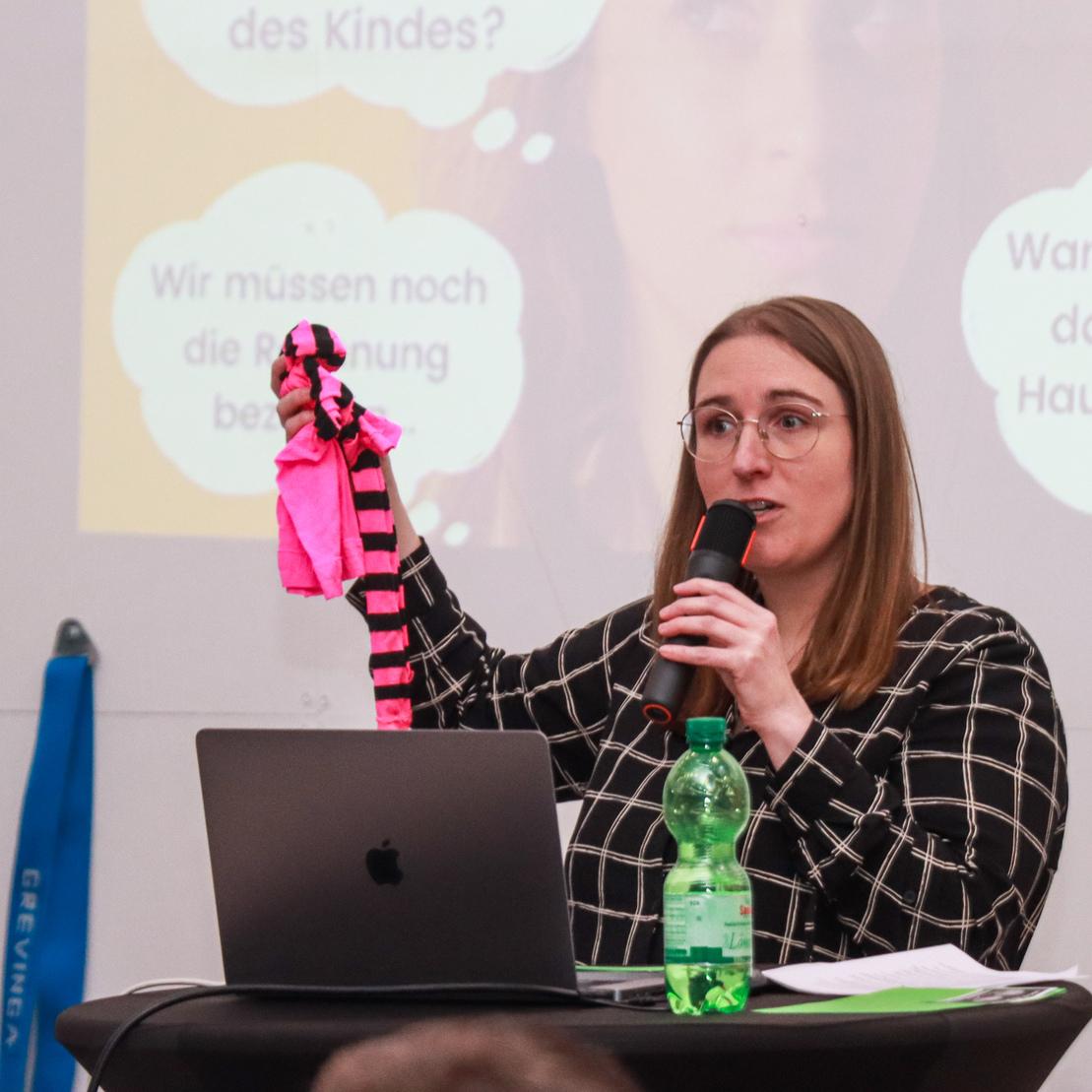 Frau spricht an einem Tisch und h&auml;lt ein pinkes, gestreiftes Stofftier w&auml;hrend einer Pr&auml;sentation.