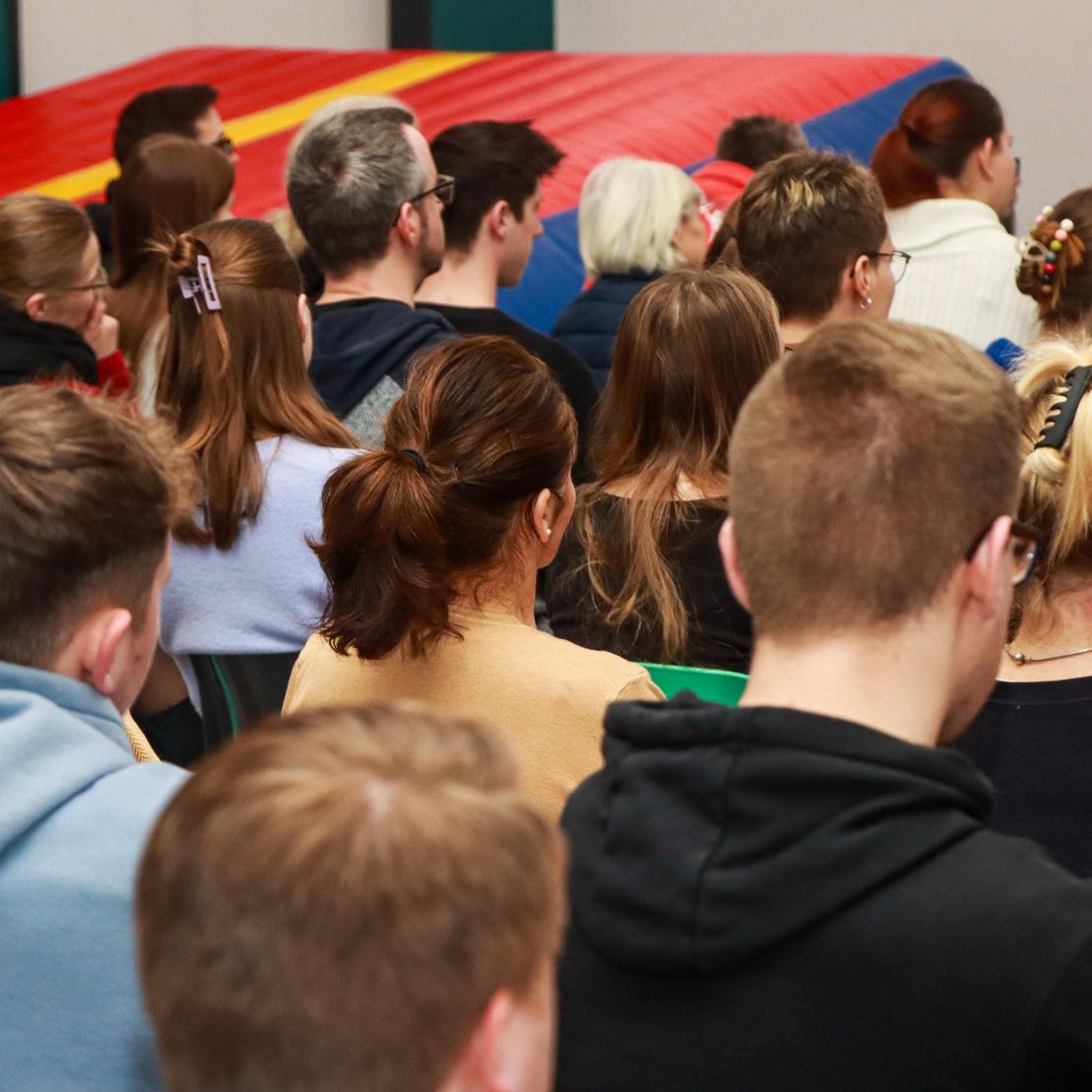 R&uuml;ckansicht einer Menschenmenge in einem Raum, mit Fokus auf die Teilnehmer und einer farbigen Turnmatte im Hintergrund.