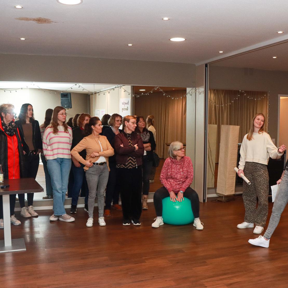 Gruppe von Frauen in einem Raum, einige stehen, andere sitzen, w&auml;hrend zwei Frauen eine Pr&auml;sentation halten.