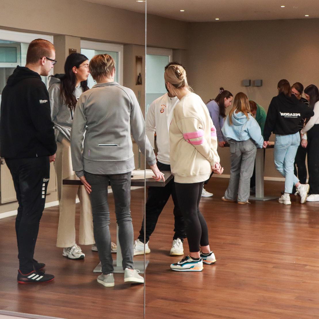 Gruppe von Menschen steht in einem Raum, einige unterhalten sich, andere arbeiten an einer Aktivit&auml;t.