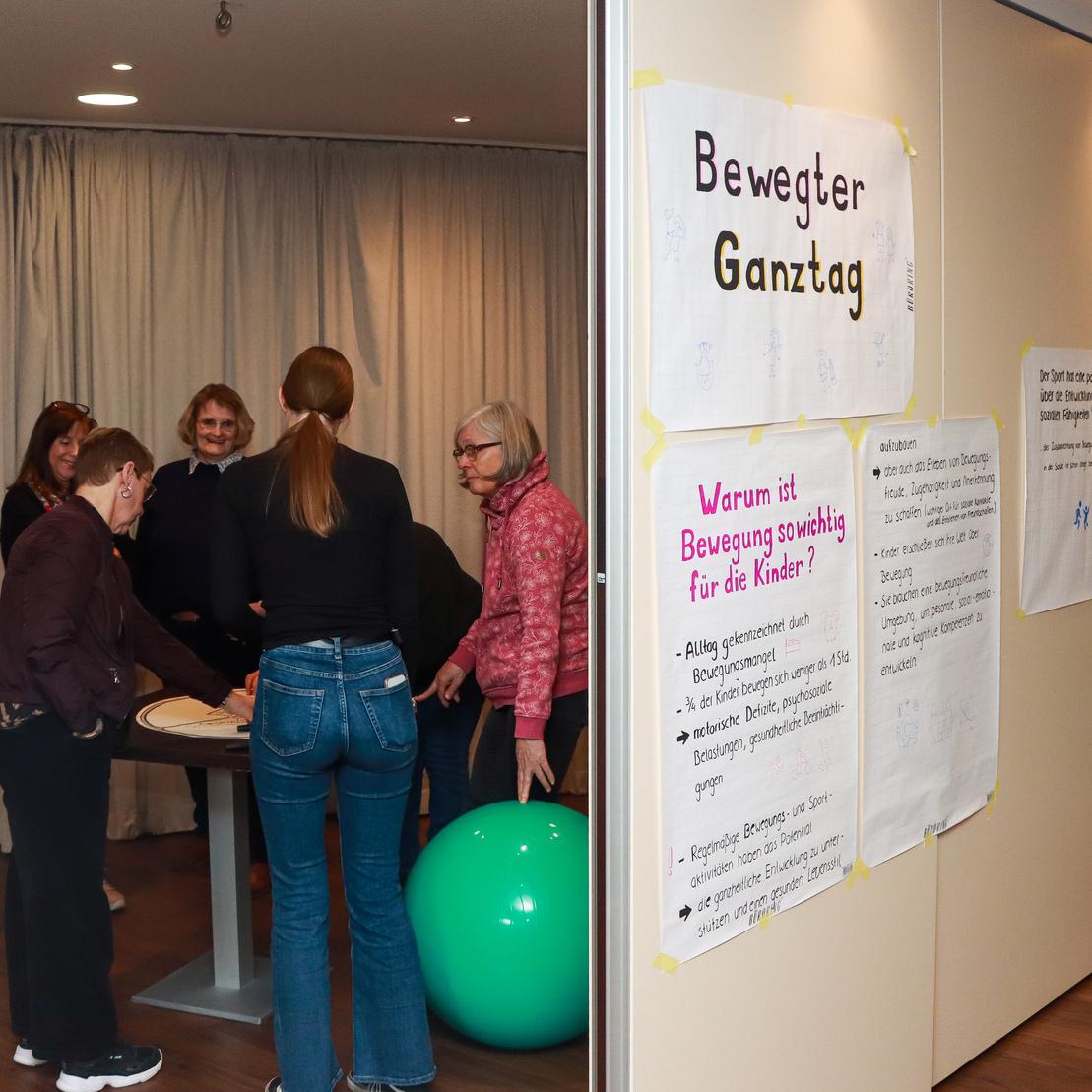Gruppe von f&uuml;nf Frauen um einen Tisch, neben einem gr&uuml;nen Gymnastikball, mit Postern zum Thema Bewegung im Hintergrund.