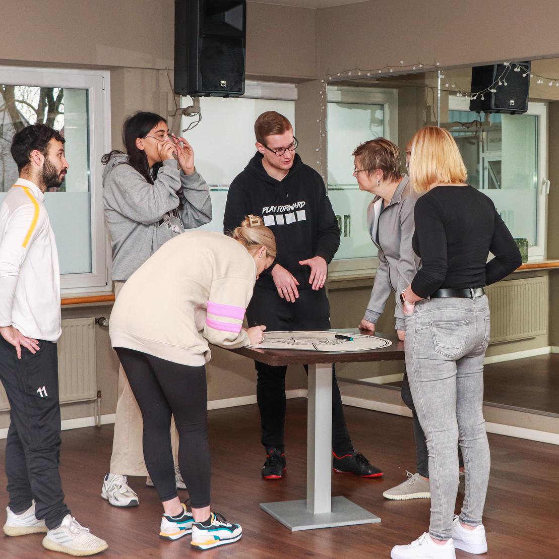 Sechs Personen stehen um einen Tisch und diskutieren, w&auml;hrend im Hintergrund ein Spiegel zu sehen ist.