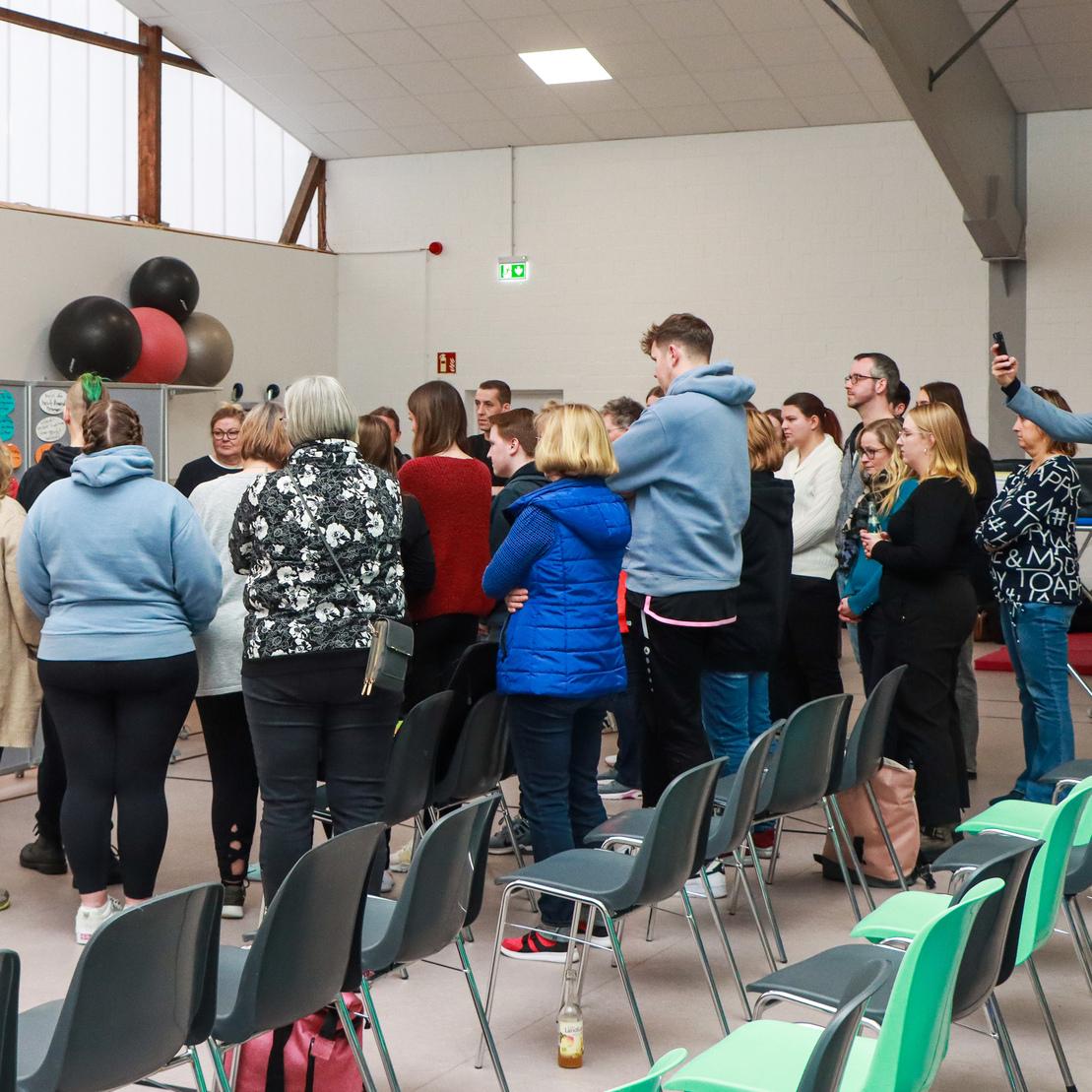 Gruppe von Personen in einem Raum, versammelt um eine Pr&auml;sentation, mit St&uuml;hlen im Vordergrund.