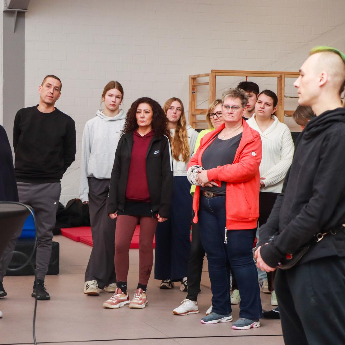 Gruppe von Menschen h&ouml;rt einer Pr&auml;sentation aufmerksam zu, w&auml;hrend eine Rednerin am Tisch steht.