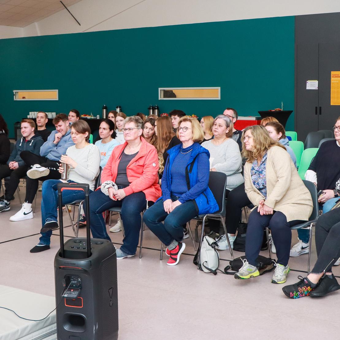 Eine Gruppe von Menschen sitzt in Reihen auf St&uuml;hlen in einem Raum mit gr&uuml;nen W&auml;nden und einem Projektor im Vordergrund.