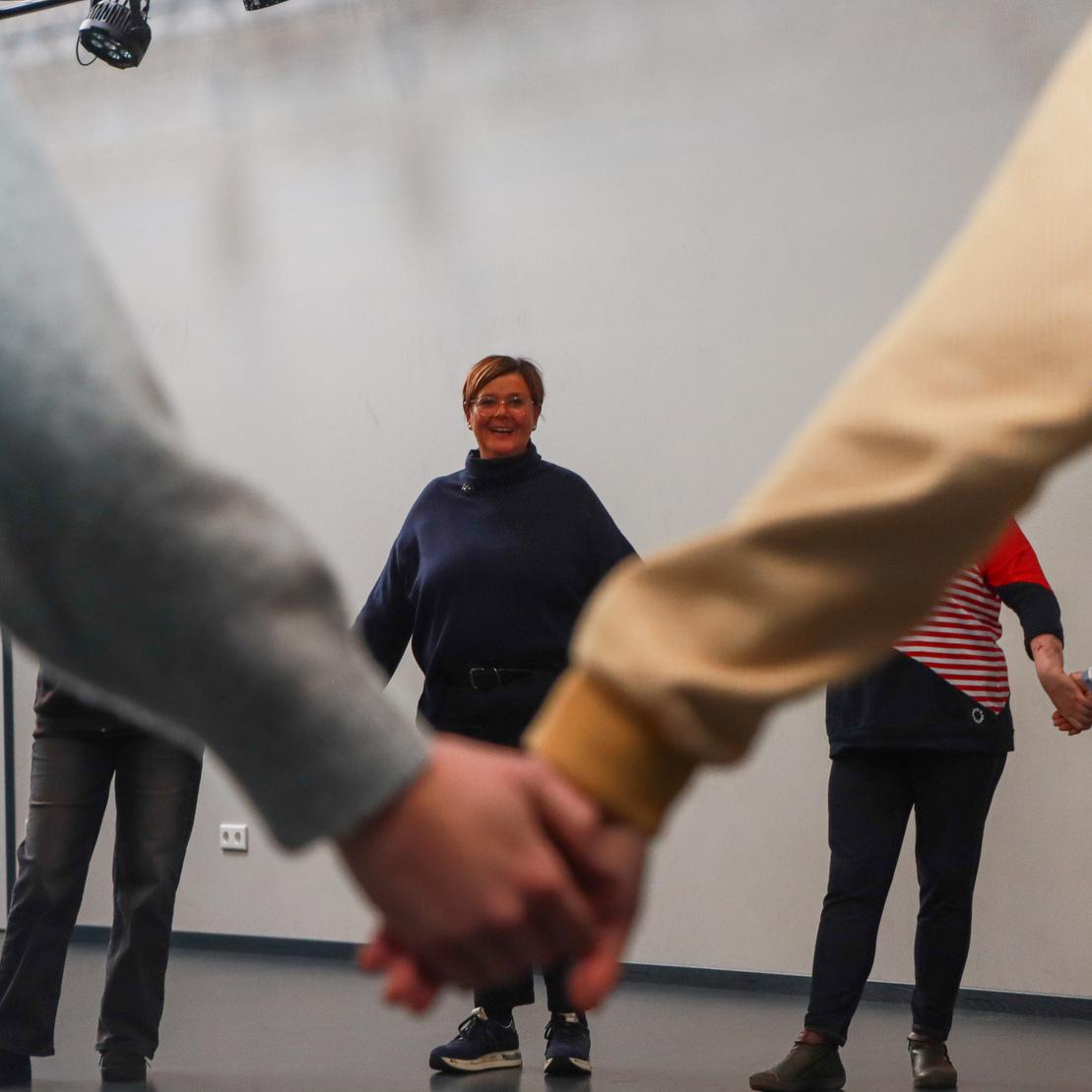 Gruppenszene mit mehreren Personen, die sich an den H&auml;nden halten, im Vordergrund eine l&auml;chelnde Frau.