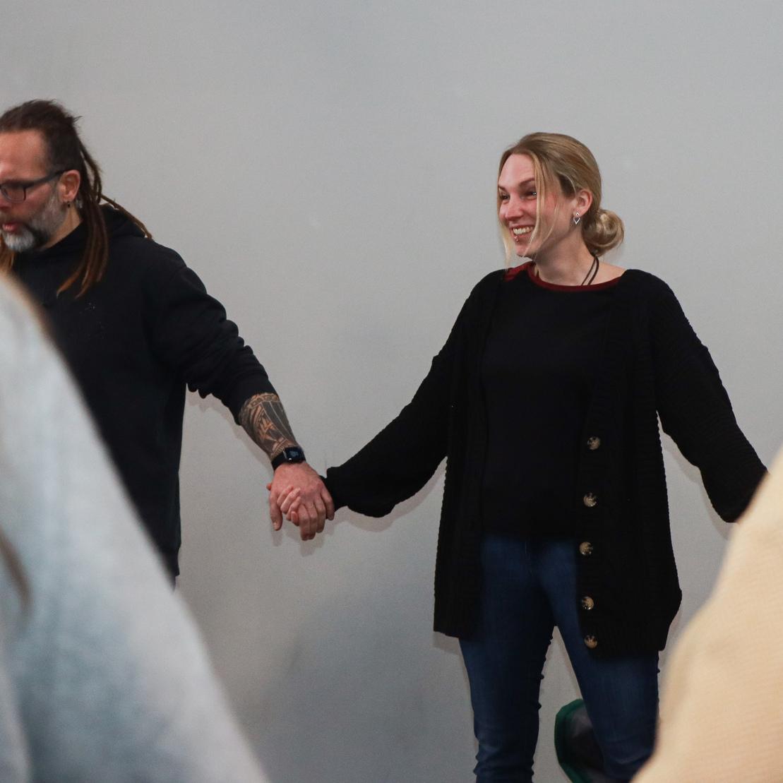 Zwei Personen stehen in einem Raum und halten sich an den H&auml;nden, w&auml;hrend sie l&auml;cheln und interagieren.