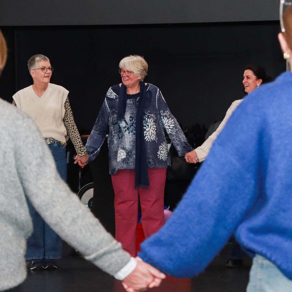 Gruppencoaching mit f&uuml;nf Personen, die sich an den H&auml;nden halten und im Kreis stehen, vor einer dunklen Wand.