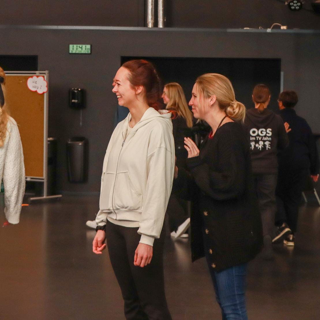Zwei Frauen in Freizeitkleidung stehen in einem Veranstaltungsraum und unterhalten sich, w&auml;hrend andere Personen im Hintergrund agieren.