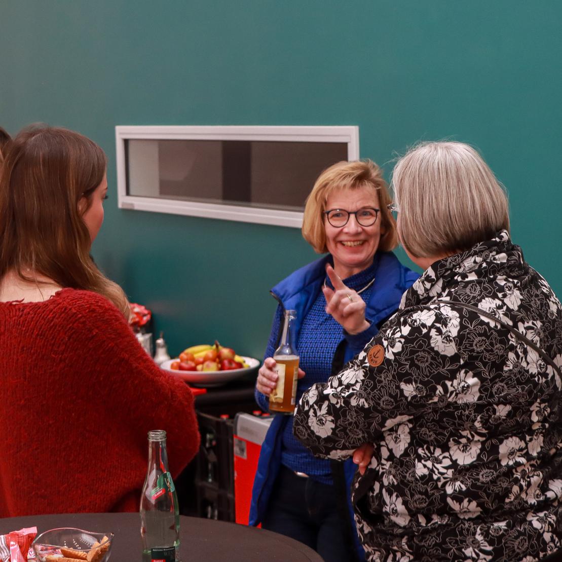 Vier Frauen unterhalten sich an einem Stehtisch, umgeben von Snacks und Getr&auml;nken, vor einer gr&uuml;nen Wand.