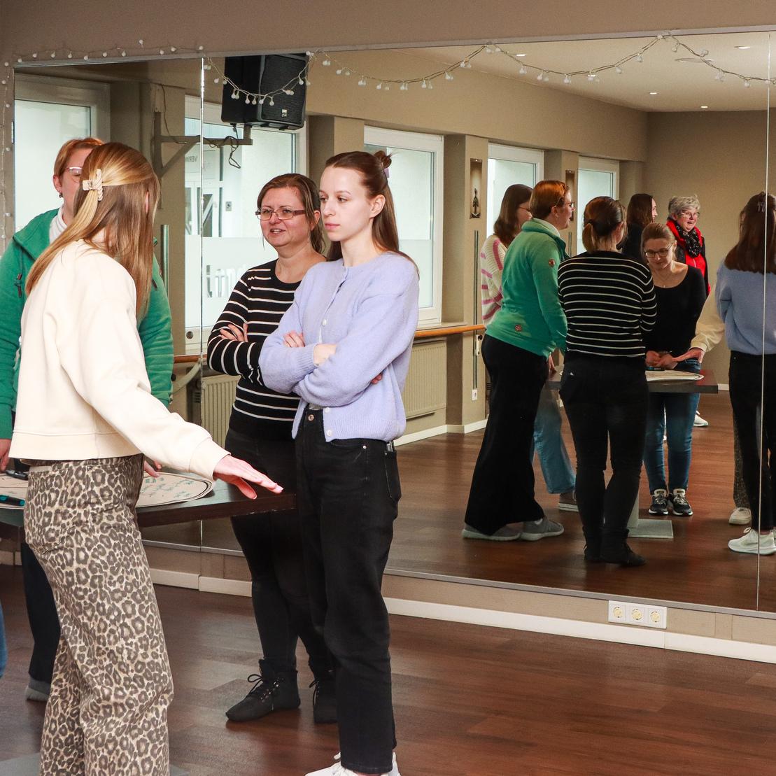 Gruppe von Frauen steht vor einem Spiegel in einem Tanzstudio und unterh&auml;lt sich. Warmes Licht und Holzfu&szlig;boden.