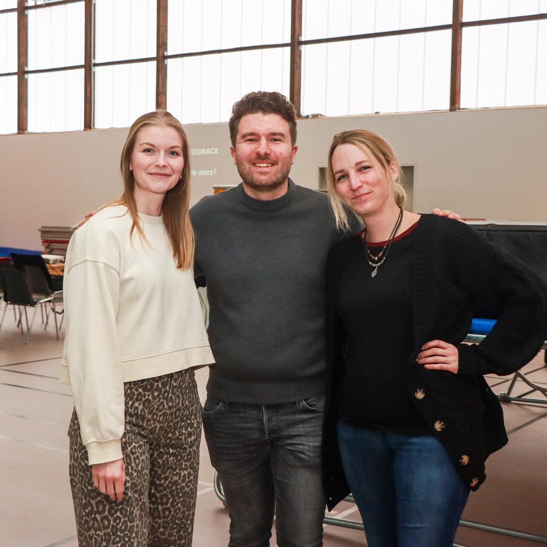 Drei Personen stehen in einer Sporthalle, umgeben von Trainingsger&auml;ten und St&uuml;hlen.