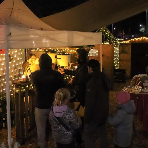 Besucher stehen an einem festlich dekorierten Stand, umgeben von Lichtern und Holzbuden auf einem Weihnachtsmarkt.
