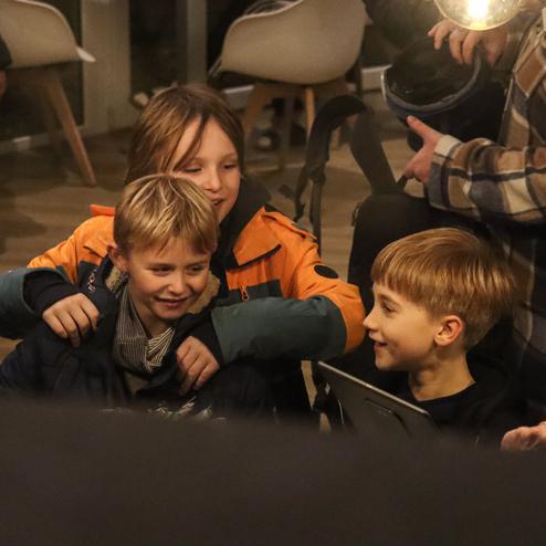 Drei Kinder sitzen zusammen und lachen, w&auml;hrend sie sich in einem hellen Innenraum unterhalten.