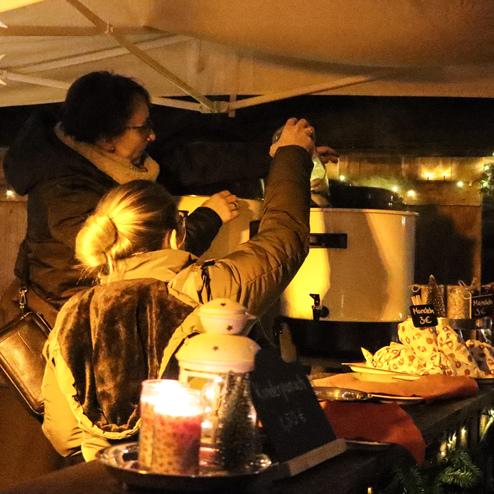 Zwei Frauen stehen unter einem Zelt und schenken Gl&uuml;hwein aus einem gro&szlig;en Beh&auml;lter aus. Weihnachtsdekoration im Hintergrund.