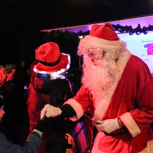 Weihnachtsmann &uuml;bergibt einem Kind ein Geschenk, umgeben von festlich gekleideten Menschen bei einer Abendveranstaltung.