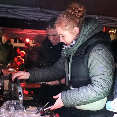 Frau serviert Speisen an einem Feststand unter einem Zelt, im Hintergrund stehen weitere Menschen.