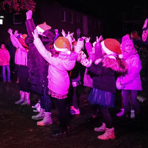 Gruppen von Kindern tragen Weihnachtsm&uuml;tzen und tanzen unter bunten Lichtern bei einer n&auml;chtlichen Veranstaltung.
