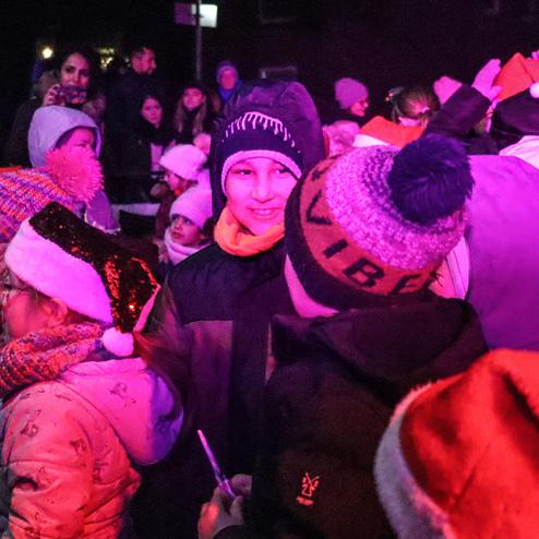 Gruppenkinder in Winterkleidung mit M&uuml;tzen und Weihnachtsm&uuml;tzen stehen in einem beleuchteten Umfeld und l&auml;cheln.