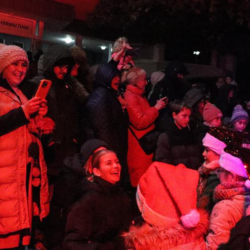 Gruppierte Menschen in winterlicher Kleidung feiern unter roten Lichtern, einige halten Smartphones, Kinder lachen.