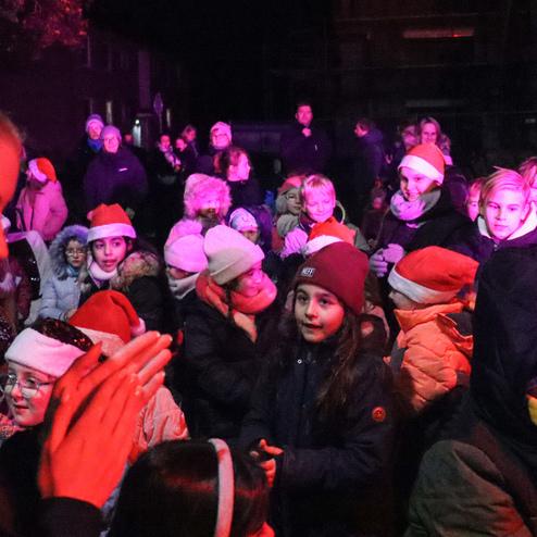 Gruppierung von Kindern in warmen Winterkleidern und Weihnachtsm&uuml;tzen, umgeben von festlichem Licht bei einer abendlichen Veranstaltung.