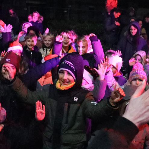 Eine Gruppe von Kindern in warmer Kleidung feiert fr&ouml;hlich bei Nacht, umgeben von bunten Lichtern.