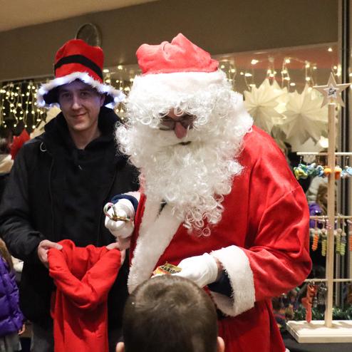 Weihnachtsmann verteilt Geschenke an Kinder, umgeben von Personen in festlicher Kleidung und bunten Deko-Elementen.