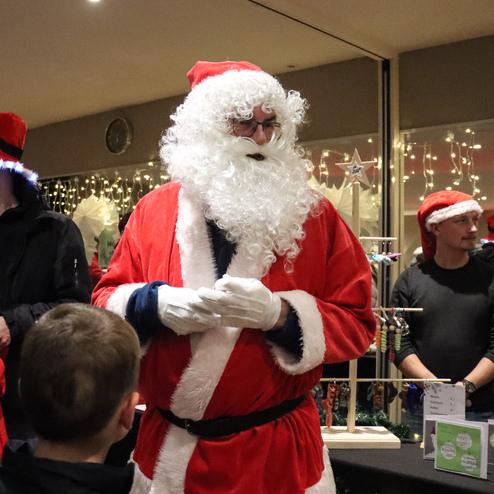 Weihnachtsmarkt mit einem Weihnachtsmann in rotem Anzug, umgeben von G&auml;sten in festlicher Kleidung und Lichtern im Hintergrund.