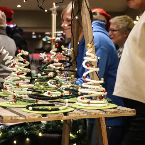 Dekorierte Weihnachtsb&auml;ume aus Draht und Filz stehen auf einem Holztisch, umgeben von Besuchern bei einem Weihnachtsmarkt.