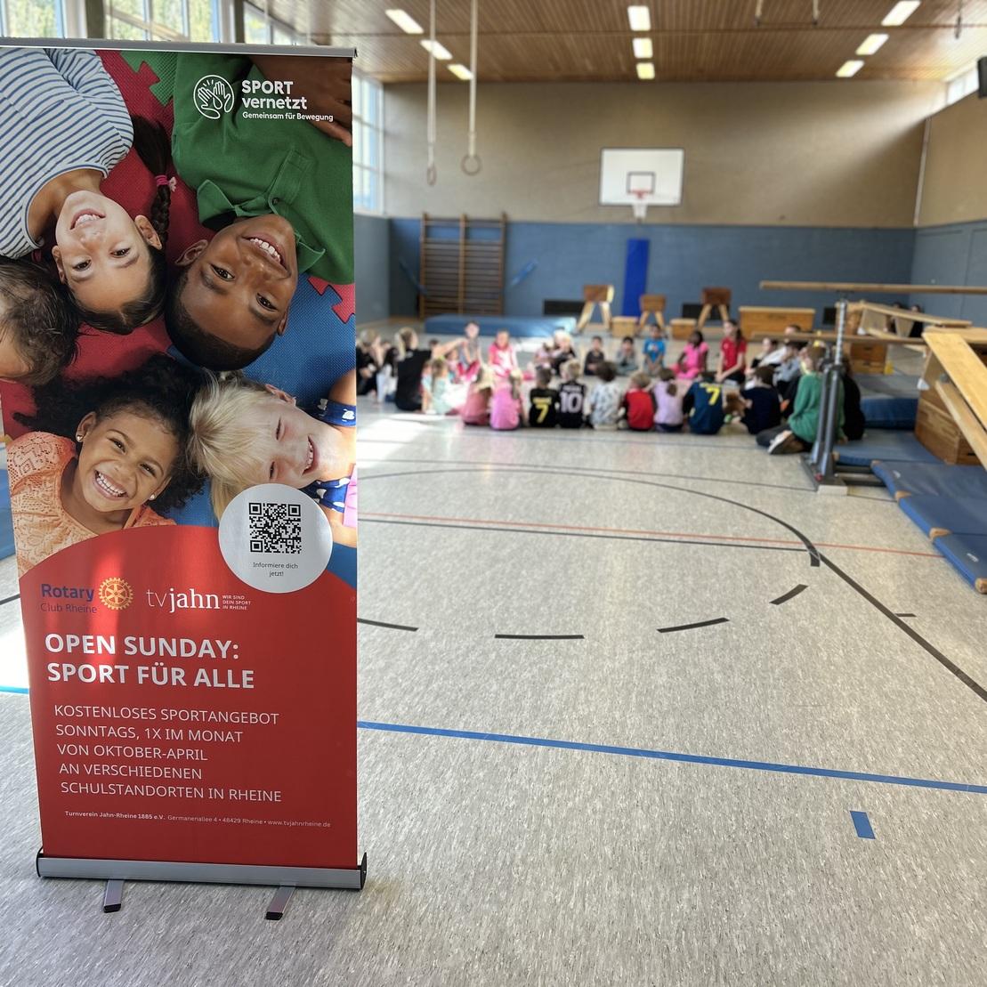 Rollup-Banner mit Kinderportr&auml;ts im Vordergrund, Hintergrund zeigt eine Sporthalle mit sitzenden Kindern.
