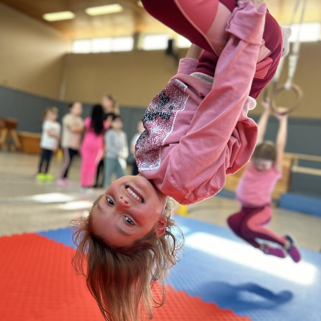 M&auml;dchen h&auml;ngt kopf&uuml;ber an einem Turnring in einer Sporthalle, l&auml;chelnd vor anderen Kindern auf einer Matte.
