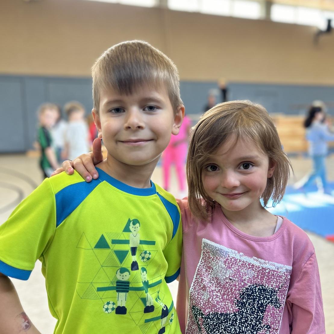 Zwei Kinder stehen l&auml;chelnd in einer Sporthalle, das Junge tr&auml;gt ein gr&uuml;nes T-Shirt, das M&auml;dchen ein rosa Oberteil.