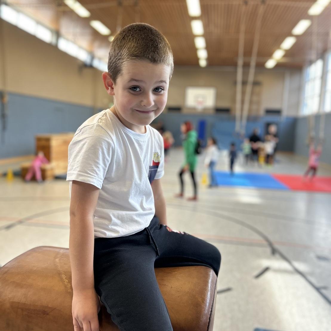 L&auml;chelnder Junge sitzt auf einem Turnblock in einer Sporthalle, w&auml;hrend andere Kinder im Hintergrund aktiv sind.